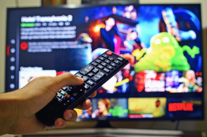 Eine Hand hält eine Fernsehfernbedienung. Im Hintergrund ist ein Fernsehbildschirm zu sehen, auf dem verschiedene Programme zur Auswahl stehen.